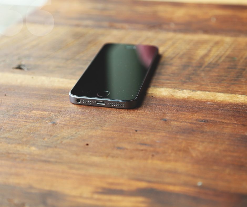 A black smartphone rests face up on a wooden table with its screen turned off. The wood’s natural grain is visible, and the phone’s charging port can be seen at the bottom.