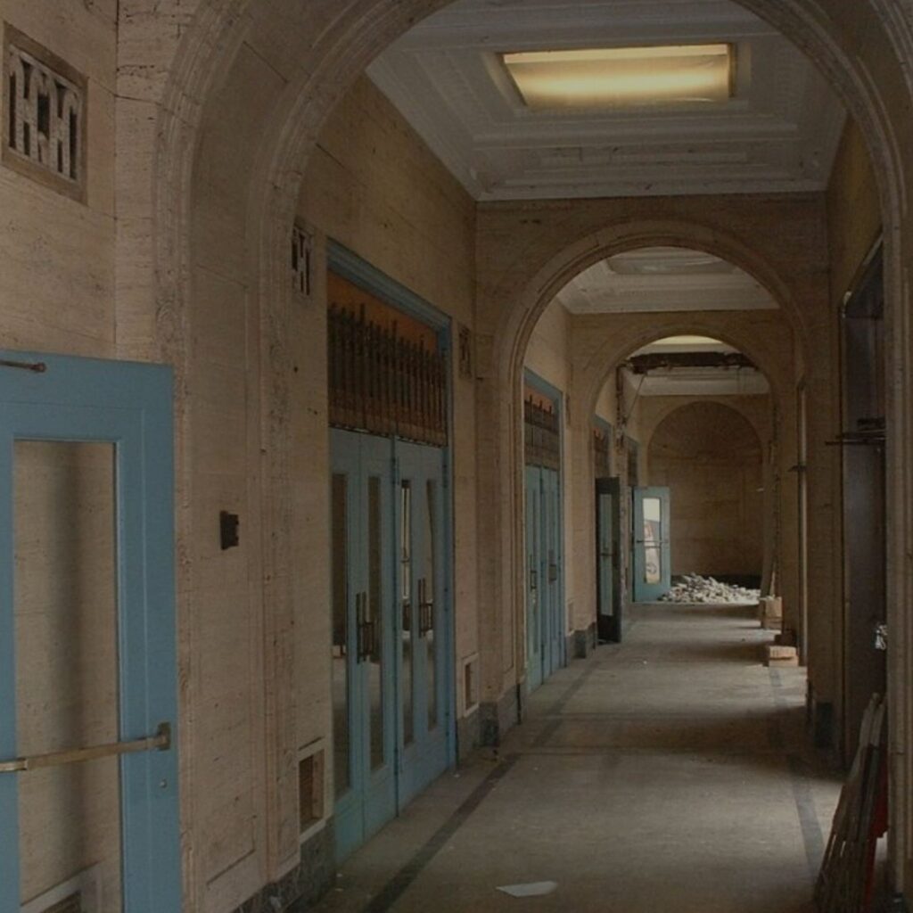 A dim hallway with blue doors, old metal handles, arched ceilings, and scattered debris at one end, suggesting the neglected state of the Philadelphia Civic Center.
