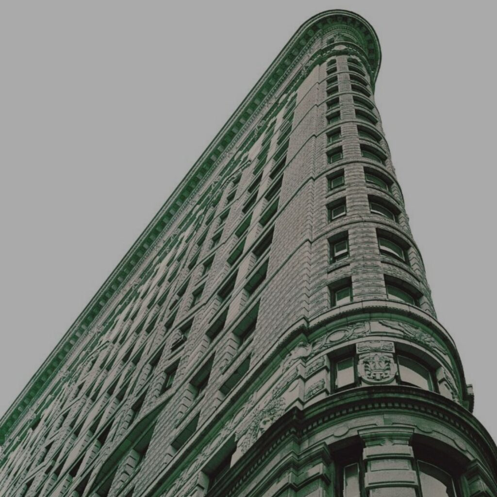 Looking up at NYC’s iconic Flatiron Building, showcasing detailed stonework and copper-trimmed windows. The triangular structure rises into an overcast gray sky.