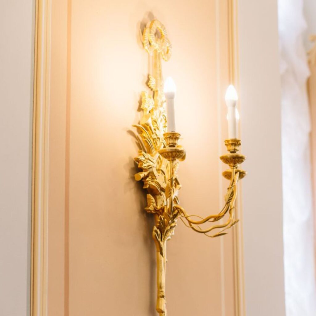 Ornate golden wall sconce with two candle-shaped bulbs, elegant and decorative, mounted on a peach wall with intricate floral designs for stylish illumination.