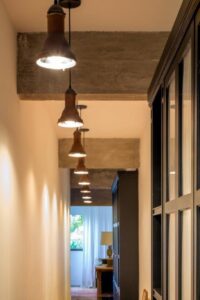 A hallway with visible bulb pendant lights and wooden beams leads to a window draped in white curtains at the end, filling the space with soft natural light.