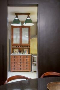 Looking through a dark doorway, you see a kitchen with green hanging lamps, a wooden cabinet, and several bottles scattered across the countertop under soft light.