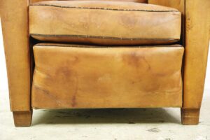 Close-up of a leather club chair showing the lower seat cushion and front legs, with visible age-related wear and stitching details.