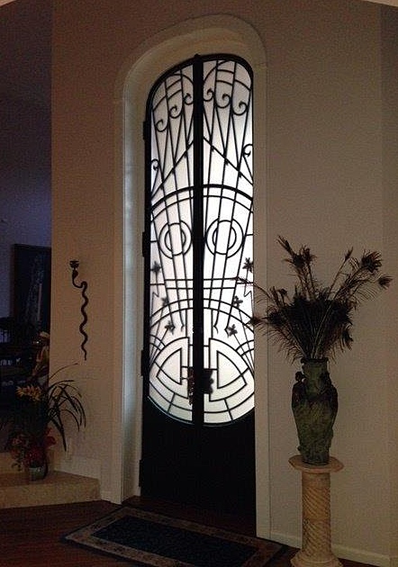 A tall, narrow door with black ironwork forms a mask-like pattern; next to it, a pedestal holds a vase filled with elegant peacock feathers.