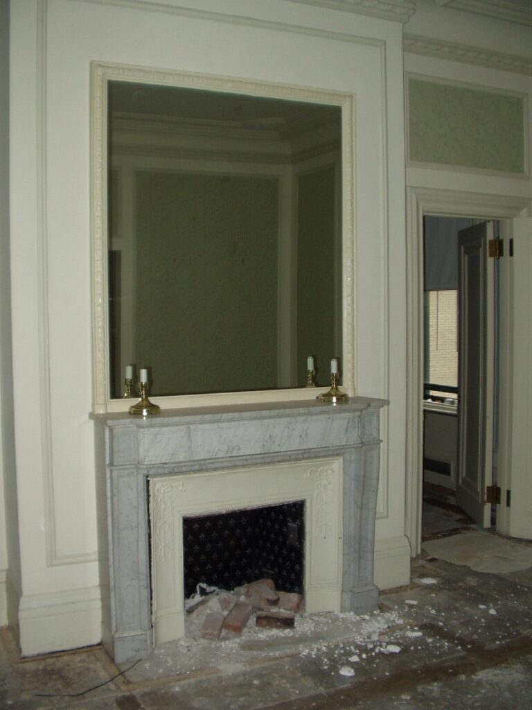 White marble mantel with large mirror above, candle holders, and bricks placed inside. A doorway adds to the scene’s Manhattan loft charm. All features are clearly visible.