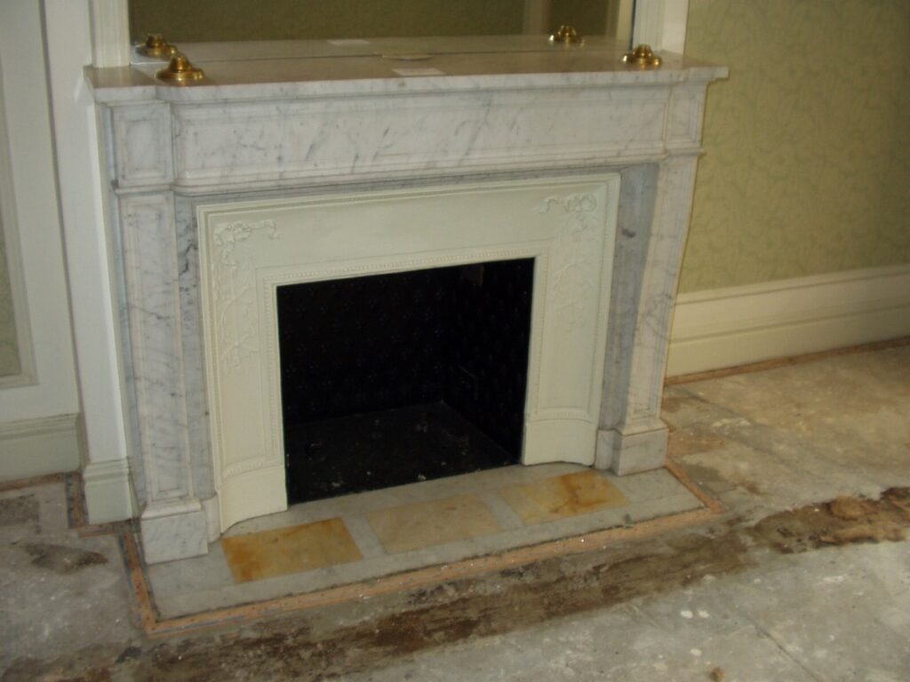 A marble fireplace with a reclaimed mantel stands against green wallpaper. The floor is in renovation, displaying exposed and unfinished patches throughout the room, indicating work in progress.