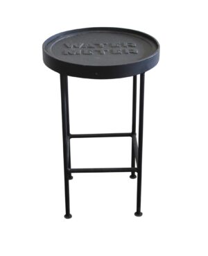 Round side table with a cast iron water meter cover top and slender metal legs, shown in a front view.