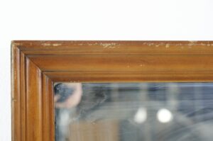 Close-up of a rectangular wall mirror with a medium wood tone frame and visible surface wear along the top corner.