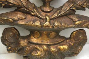 Close-up of an antique bronze applique with an urn shape and ornate leaf details, showing the mounting holes and central band.