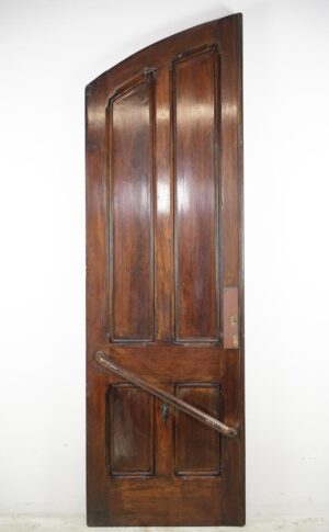 Antique wooden double door with an arched top, four vertical panels, and a diagonal brace, shown in dark wood tone with visible wear near the bottom.