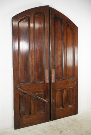Antique double doors with an arched top and four vertical panels each, shown closed and made of dark wood tone wood, with visible wear near the bottom edges.