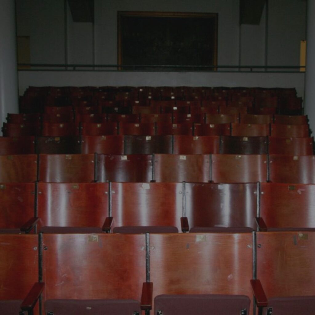 Rows of glossy wooden auditorium seats face forward in a dimly lit Great Hall. The darkened background emphasizes the empty, orderly rows, creating a quiet and spacious atmosphere.