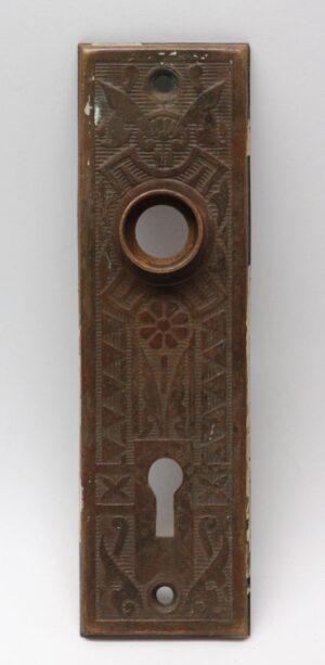 Vertical stamped brass door back plate with an antique brass finish, featuring a round knob opening, keyhole cutout, and decorative embossed pattern, shown front view with visible wear.