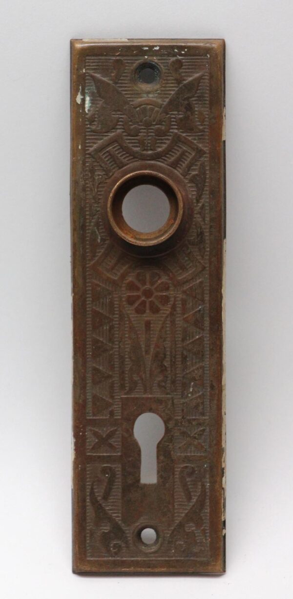Vertical stamped brass door back plate with an antique brass finish, featuring a round knob opening, keyhole cutout, and decorative embossed pattern, shown front view with visible wear.