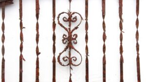 Heavily rusted wrought iron fire escape balcony front with ornate scrollwork and vertical bars, showing chipped paint and visible bends in the frame.