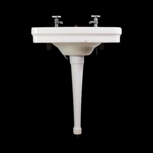 1920s white porcelain pedestal sink with two taps and a long tapered leg, shown front on with visible mounting hardware beneath the basin.