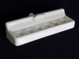 White ceramic wall-mount toothbrush holder with six brush slots, shown in a close-up angled view with visible surface wear and staining.