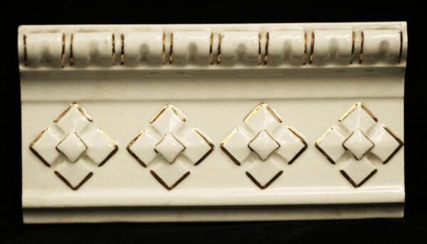 Off-white ceramic trim tile with a gold geometric pattern and molded top edge, shown in close-up.