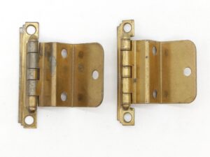 Pair of semi concealed cabinet hinges shown side by side, each with an offset barrel and mounting holes, finished in antique brass with visible surface wear.