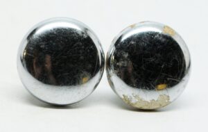 Pair of chrome cabinet knobs shown side by side, each round with visible age-related surface wear and one knob showing more tarnish than the other.