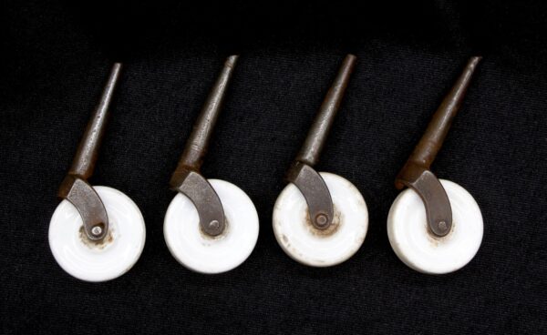 Four antique white porcelain caster wheels with tapered steel stems are shown in a close-up, arranged in a row on a dark background.