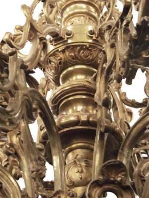 Close-up of a multi-tiered bronze chandelier with ornate scrolling arms, decorative bobeches, and candle-style sockets.