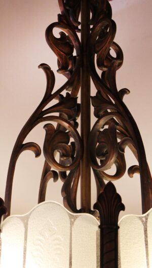 Close-up of an Art Deco chandelier with an antique bronze frame and frosted glass panels, showing the ornate scrollwork and vertical central stem.