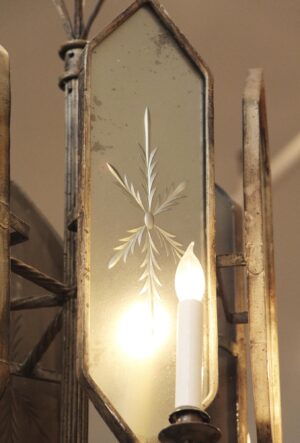 Close-up of an Art Deco chandelier panel with etched mirrored glass and an antique brass frame, showing one lit candle-style bulb and part of the wrought iron structure.