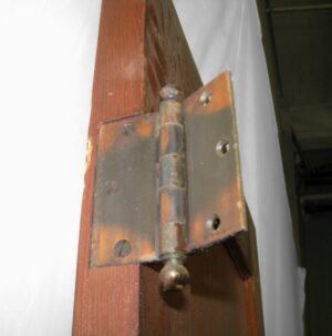 Close-up of a metal door hinge mounted on a narrow pine closet door, showing the edge of the door and hinge hardware.