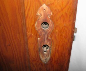 Close-up of a narrow pine closet door showing an ornate keyhole escutcheon and lock opening on the dark wood tone side.