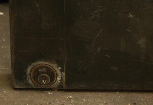 Close-up of a black metal door showing the lower corner with a round lock and visible wear on the panel.
