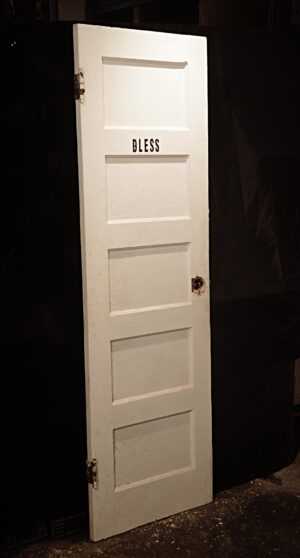 Vintage white wooden passage door with five panels, shown at an angle and marked BLESS, with hinges and a knob hole visible.