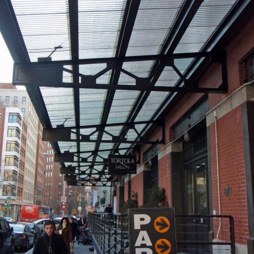 Pedestrians stroll beneath striking metal and glass canopies by urban buildings, passing signs for Tortola Salon and Parking that brighten the busy, active city sidewalk scene.