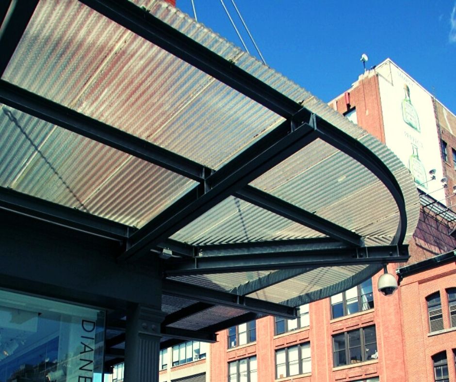 A reclaimed corrugated glass awning with a black steel frame extends from the building. Behind it are brick structures beneath a clear blue sky, offering an urban architectural scene.