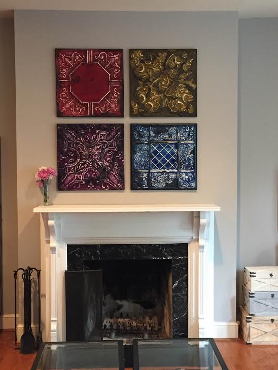 Our tin looks amazing over a mantel A white Olde Good Things fireplace stands against a gray wall, decorated with pink flowers in a vase and four square art pieces arranged above. The scene is simple and elegant.