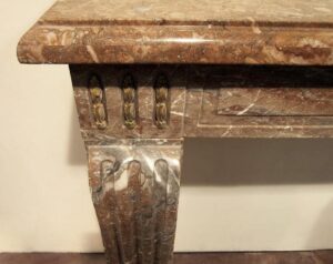 Close-up of a marble mantel with antique bronze detail appliques on the face and a shelf above, showing the left front corner and decorative side support.