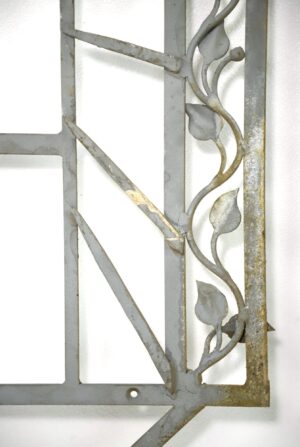 Gray wrought iron wall accent with an arched vine and leaf design, shown in close-up with light rust along the frame and edges.