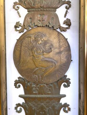 Antique bronze ornament with a central relief of the goddess Terina, surrounded by decorative scrollwork and urn details in a vertical panel.