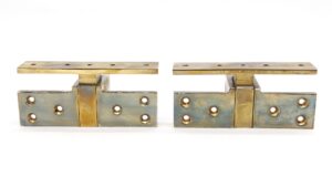 Pair of polished brass pocketed pivot door hinges shown side by side, each with a long top plate and multiple screw holes, with visible age-related wear.