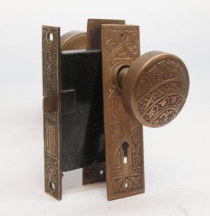 Antique passage door set with two ornate bronze knobs, matching back plates, and a black mortise lock body shown in a three-quarter view.