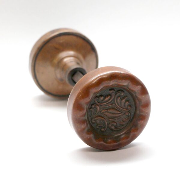 Pair of antique bronze passage door knobs shown in close-up, with one knob in front displaying an ornate Rococo medallion design and the other blurred behind it.