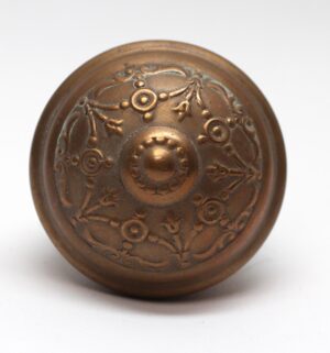 Antique brass passage door knob with a round Victorian floral and scroll relief pattern, shown front-on.