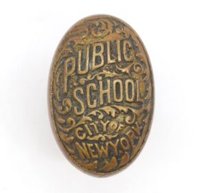 Oval antique bronze door knob with raised Public School and City of New York lettering and ornate scrollwork.