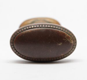 Close-up of an antique brass oval door knob with beaded edge detail and visible wear, shown from the front with a second knob blurred behind it.