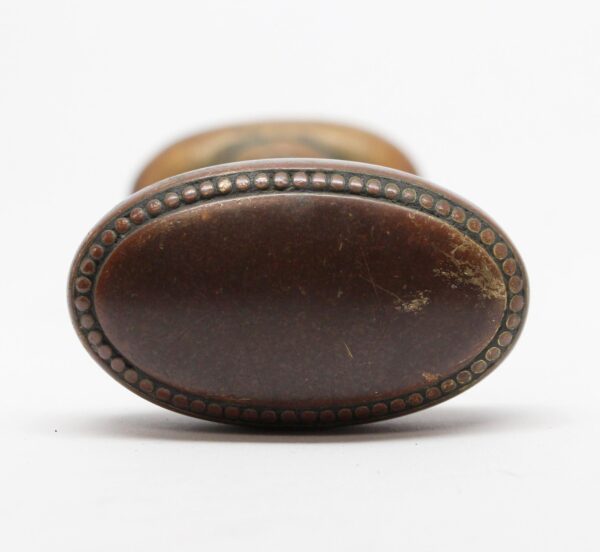Close-up of an antique brass oval door knob with beaded edge detail and visible wear, shown from the front with a second knob blurred behind it.