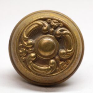 An antique brass door knob with an ornate circular face and raised scrollwork design, shown in close-up.