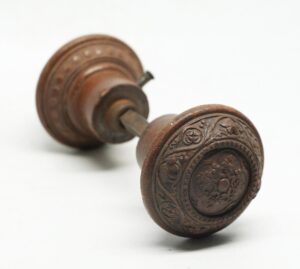 A pair of Victorian wood door knobs with a spindle, showing one knob in focus with concentric carved rings and a beaded floral scroll pattern.