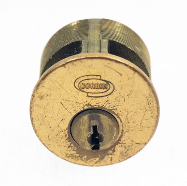 Front view of a Corbin lock cylinder with a keyway and worn antique brass finish, shown without keys.