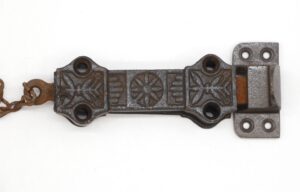 Victorian cast iron surface door bolt with spring-loaded latch and attached chain, shown closed with decorative embossed plates and mounting holes visible.
