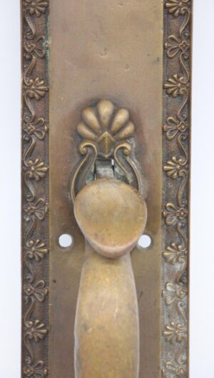 Close-up of a neoclassical bronze door pull with a flower and shell design, showing the thumb latch, lock insert, and bent plate.